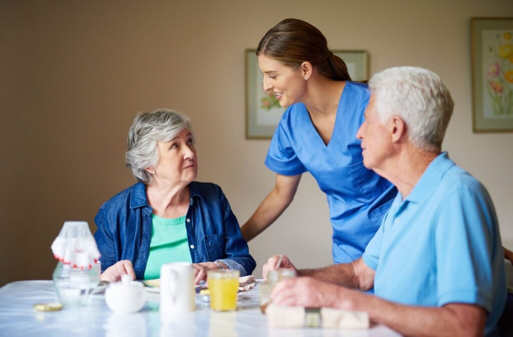 A smiling caregiver checks in on two residents during a quick meal in senior living