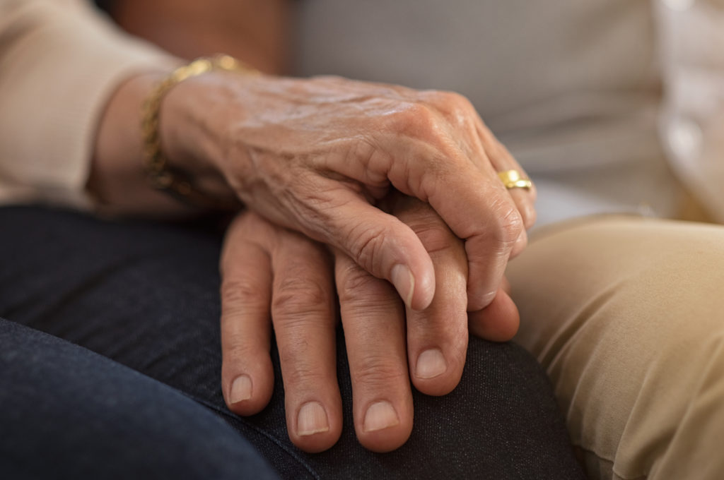 Closeup of elderly couple holding hands while sitting on couch. Husband and wife holding hands and comforting each other. Love and care concept.