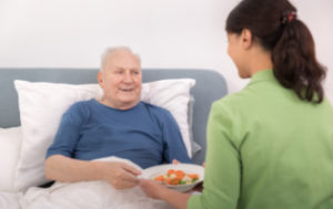 A woman handing a meal to a senior man lying in bed