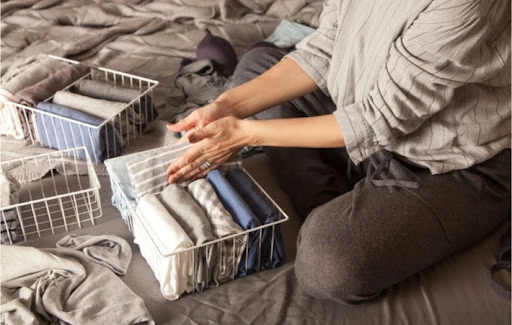A woman sitting crosslegged on a bed folding and putting away clothing into wire organizational baskets.