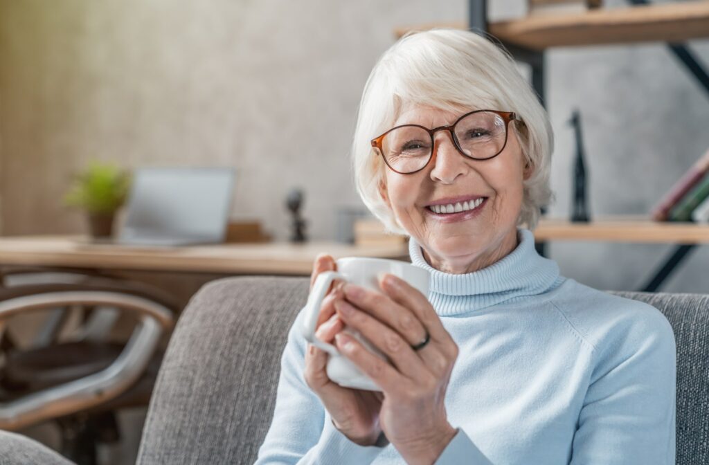 An older woman holding a cup of coffee in her hands while she sits on her couch, smiling at the camera