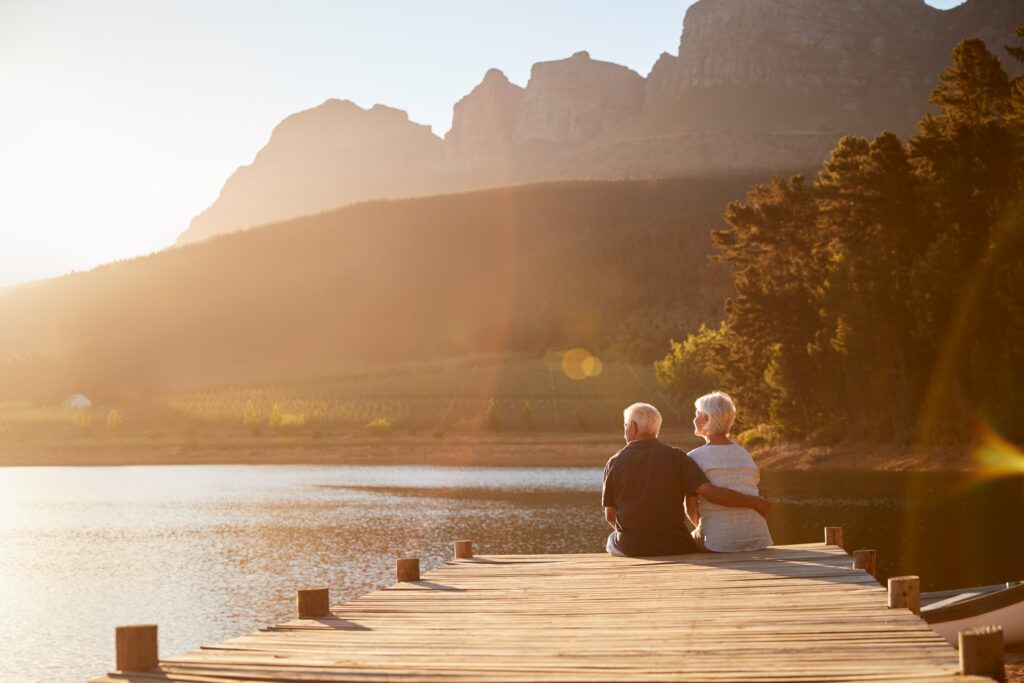A senior couple sitting at the end of a boat dock on a lake, looking at the sunset
