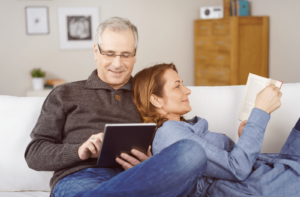 A middle-aged couple relaxing together at home with the wife lying back against her husband as she reads a book while he surfs the internet on a tablet