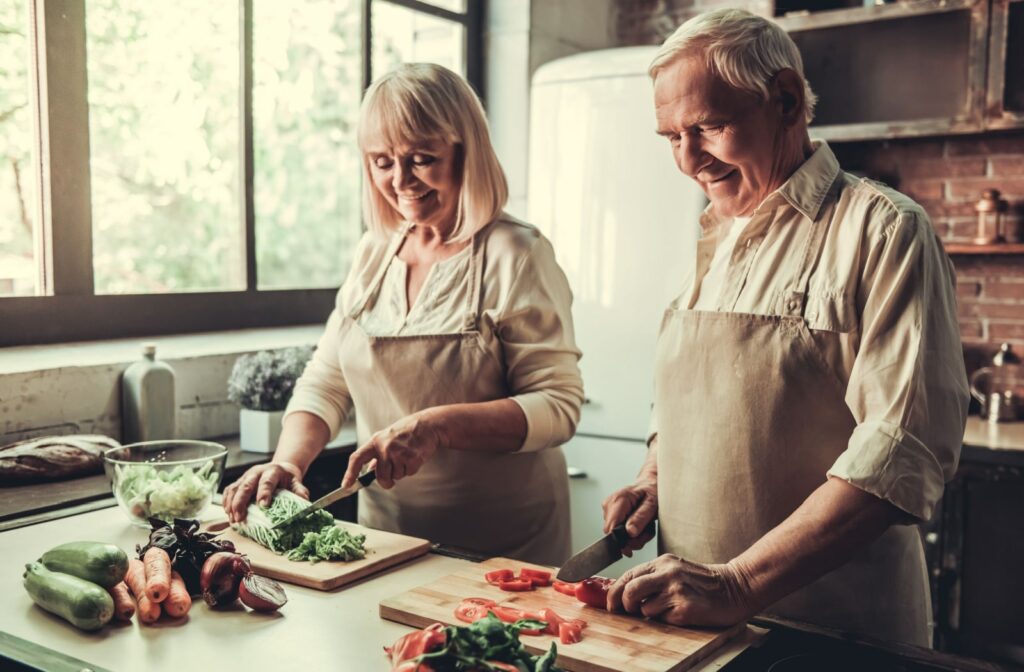 A senior couple in aprons is slicing vegetables for salad