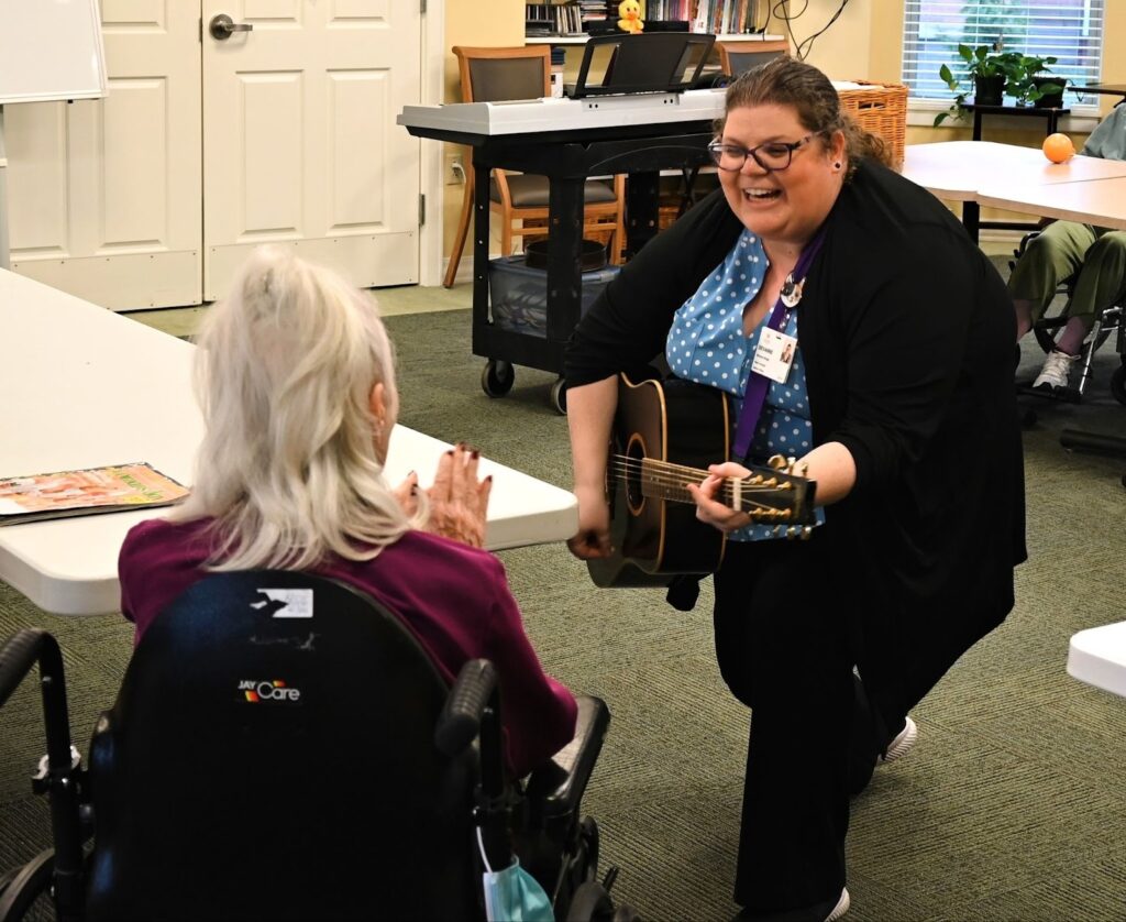 A music therapist kneels to play guitar close to a senior in a wheelchair, who claps along in enjoyment