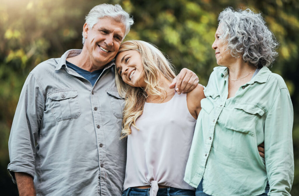 A senior sits on their bed beside their adult child, opening a gift. The senior's face is lit up with joy, and their child has their arms around their parent's shoulders in a loving manner.