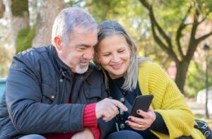 Two older adults sitting on a bench outside and scrolling Facebook on a phone.