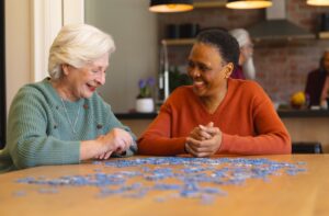 Two seniors sitting at a table in a care home participating in a puzzle while smiling and laughing together.
