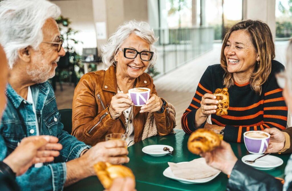 A happy group of seniors sitting around a table enjoying coffees and pastries with bright purple dishware while laughing and smiling
