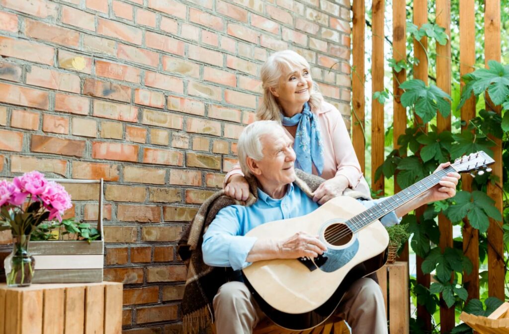 A senior couple on a bright day sitting in an outdoor gathering area in a senior living community playing guitar and smiling