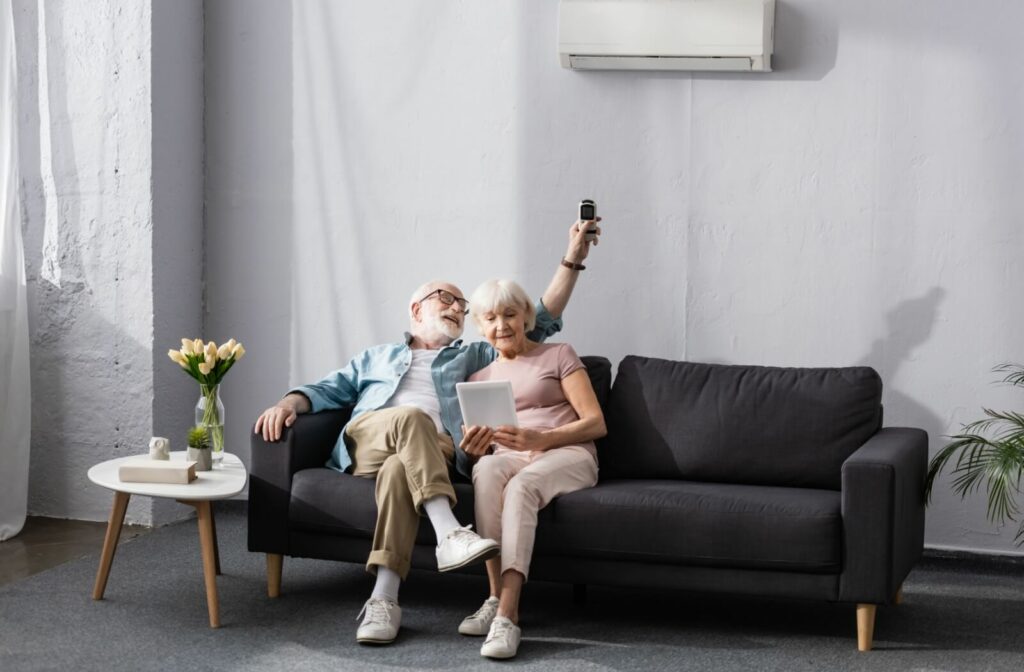 A senior couple relax on a couch in their assisted living home, adjusting the AC to their liking to stay cool in the summer
