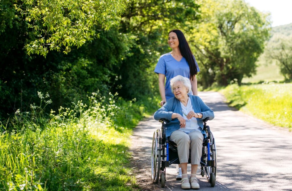 A carer pushes a senior woman in a wheelchair on a paved sidewalk in a wooded area