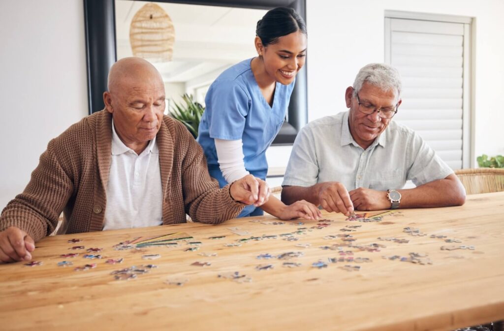 A caregiver in memory care helps two residents assemble a jigsaw puzzle on a large wooden community table