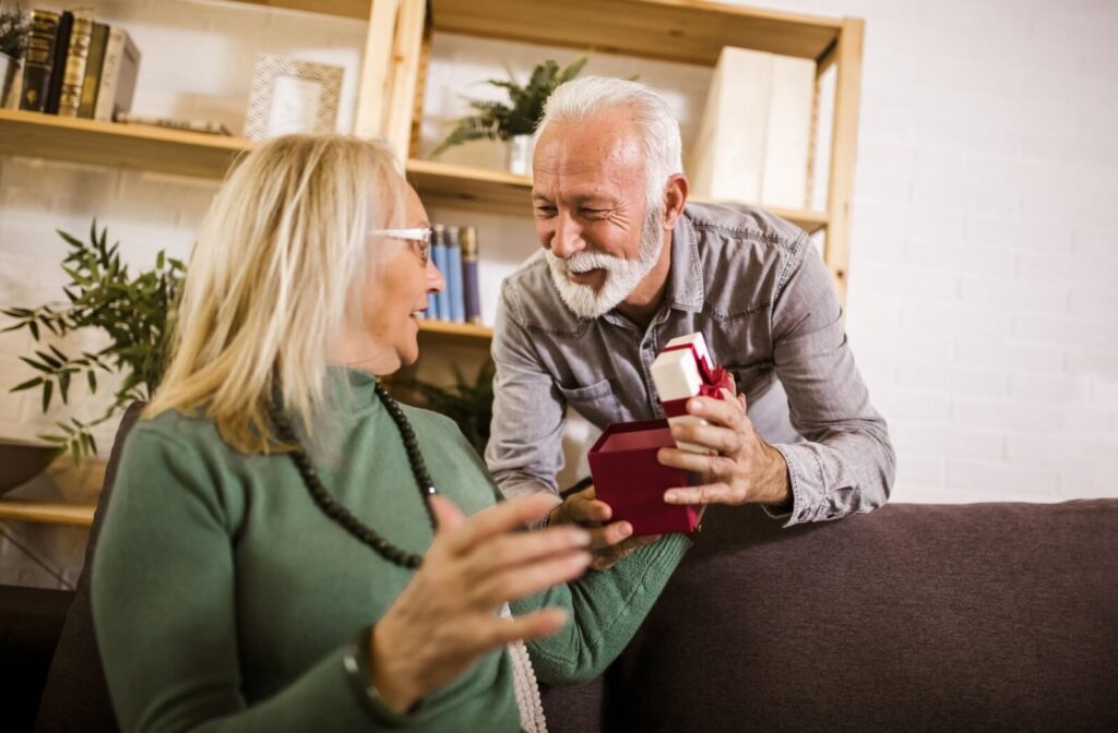 An older adult smiles while handing their seated spouse a well-wrapped holiday present