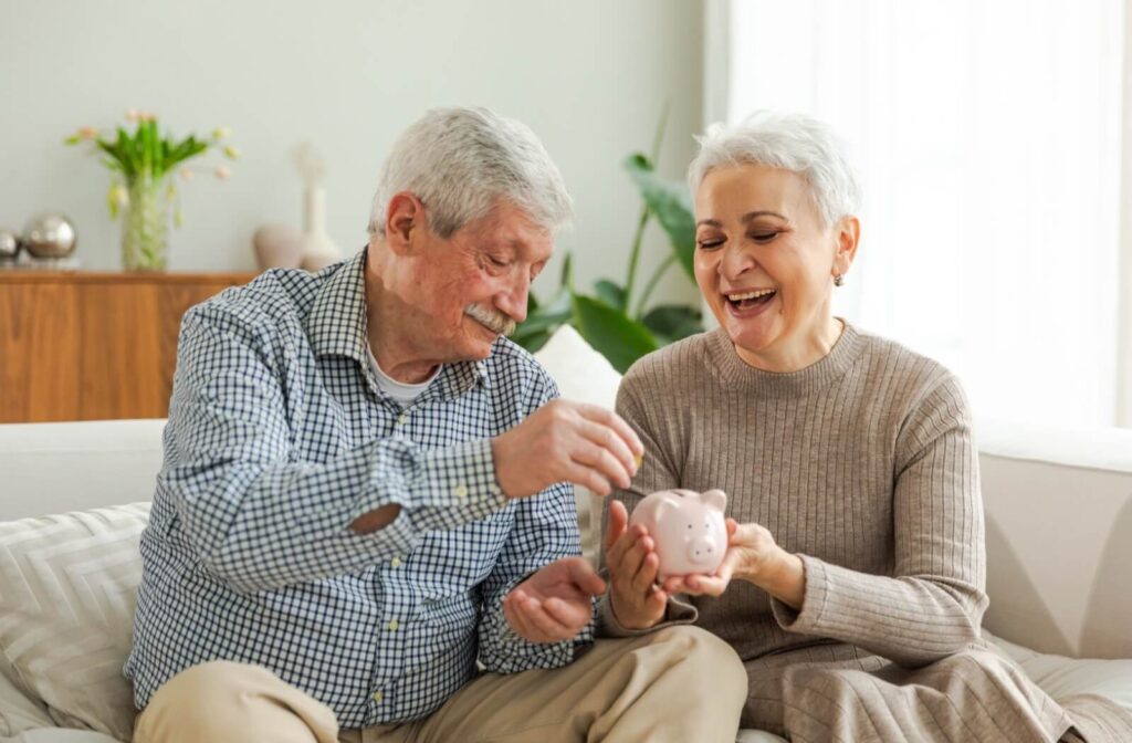 An older adult puts a coin into a piggy bank while their spouse laughs while they're sitting on a bed together