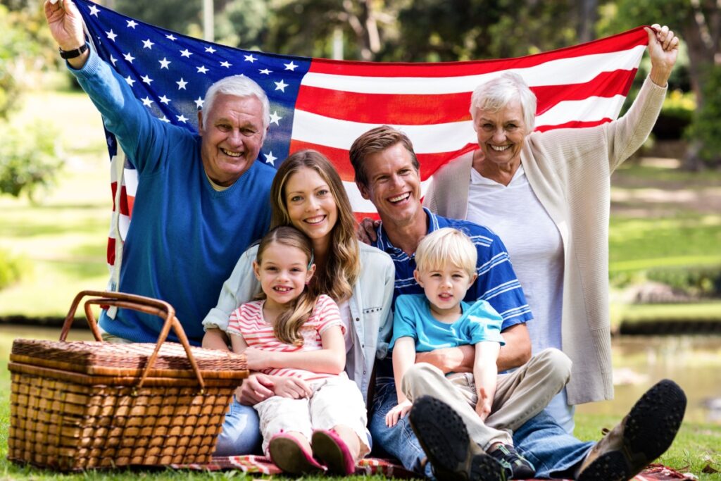 A senior couple happily holding the American Flag behind them while seated in a park with their children and grandchildren